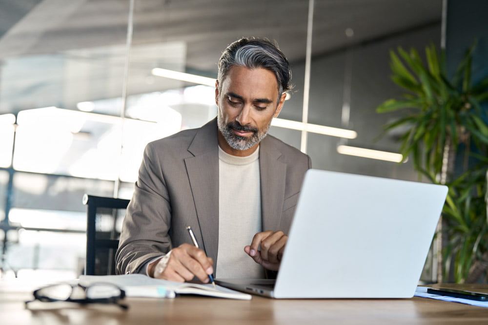 Busy mature business man writing watching webinar working in office at desk.